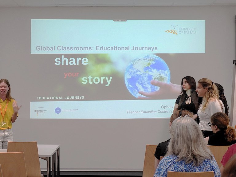 Vier Studierende stehen vor einer Gruppe Zuh&ouml;rerinnen und Zuh&ouml;rern. Einer der Studierenden spricht. Im Hintergrund ist das Titelbild einer PowerPoint-Pr&auml;sentation zum Thema "Global Classrooms: Educational Journeys" zu sehen.