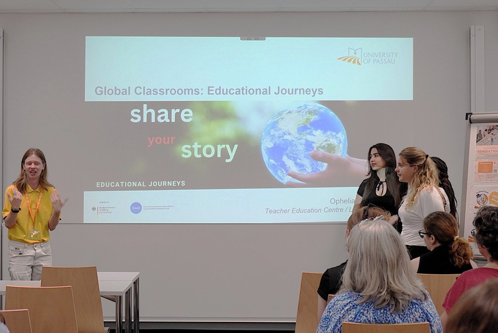 Vier Studierende stehen vor einer Gruppe Zuh&ouml;rerinnen und Zuh&ouml;rern. Einer der Studierenden spricht. Im Hintergrund ist das Titelbild einer PowerPoint-Pr&auml;sentation zum Thema "Global Classrooms: Educational Journeys" zu sehen.