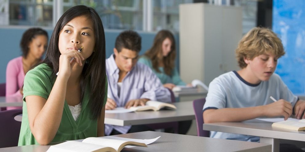 Eine Schülerin sitzt in einem Klassenzimmer und schaut nachdenklich zur Seite. Im Hintergrund sind weitere Schüler zu sehen.