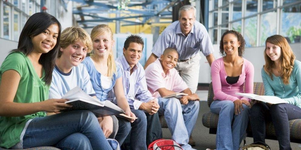Studierende sitzen in einem Halbkreis zusammen. Der Lehrer steht im Hintergrund. Alle gucken in die Kamera.