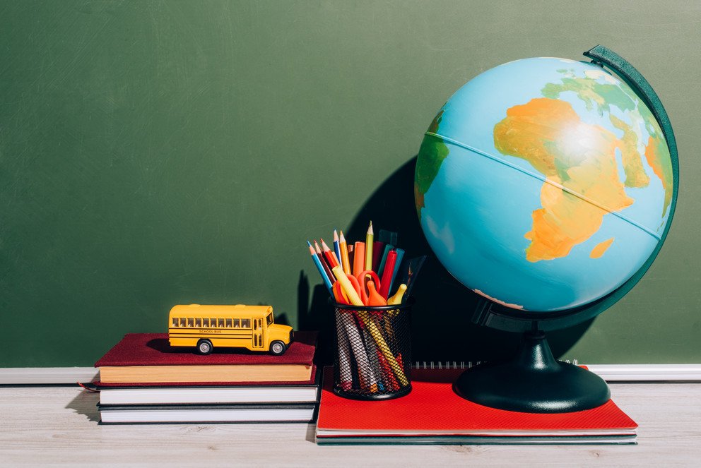 Ein Globus, ein Stfitehalter und ein Stapel aus drei B&uuml;chern, darauf ein Modell-Schulbus stehen vor einer gr&uuml;nen Schultafel