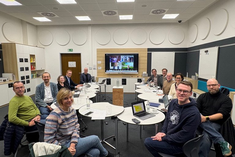 Treffen der bayerischen Zentren f&uuml;r Lehrkr&auml;ftebildung ist abgebildet. Die Beteiligten sitzen im Didaktischen Innovationslabor der Universit&auml;t Passau im Kreis um einen Tisch herum und l&auml;cheln in die Kamera.