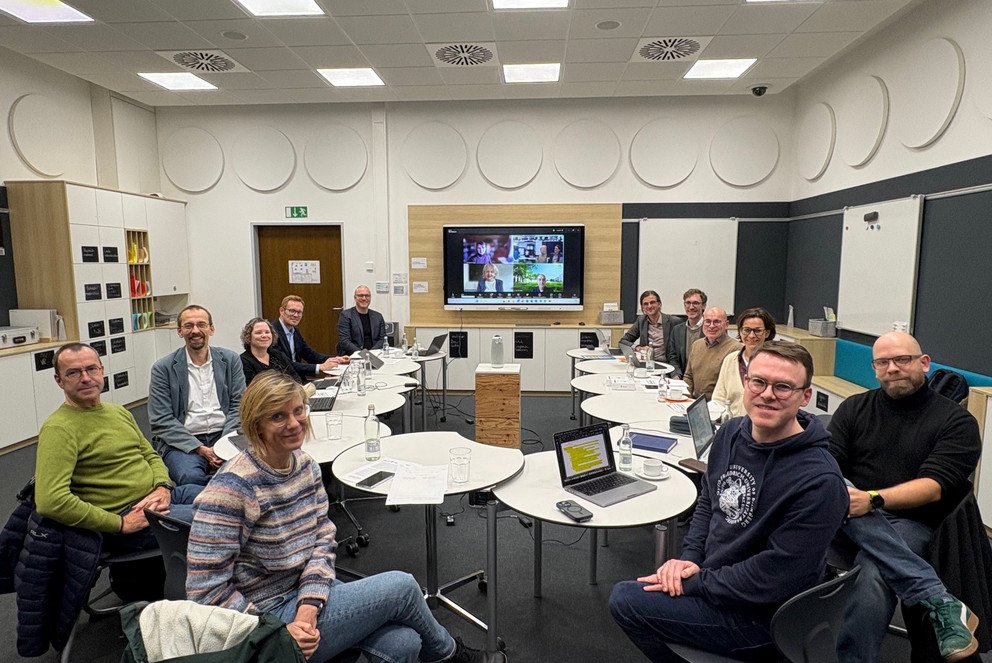 Treffen der bayerischen Zentren f&uuml;r Lehrkr&auml;ftebildung ist abgebildet. Die Beteiligten sitzen im Didaktischen Innovationslabor der Universit&auml;t Passau im Kreis um einen Tisch herum und l&auml;cheln in die Kamera.