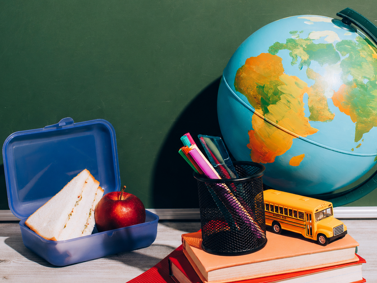Schreibtisch, auf dem ein Globus, eine gef&uuml;llte Brotdose, ein kleiner B&uuml;cherstapel, ein Becher mit Stiften und ein Modell-Schulbus stehen