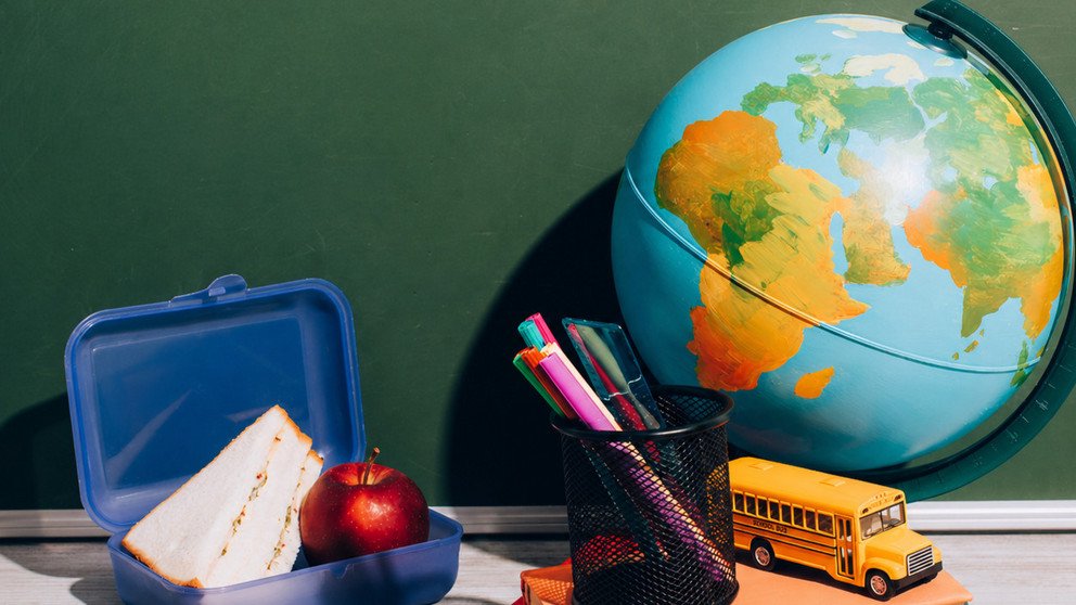 Schreibtisch, auf dem ein Globus, eine gef&uuml;llte Brotdose, ein kleiner B&uuml;cherstapel, ein Becher mit Stiften und ein Modell-Schulbus stehen