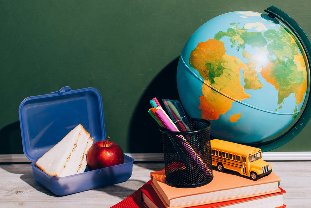 Schreibtisch, auf dem ein Globus, eine gef&uuml;llte Brotdose, ein kleiner B&uuml;cherstapel, ein Becher mit Stiften und ein Modell-Schulbus stehen