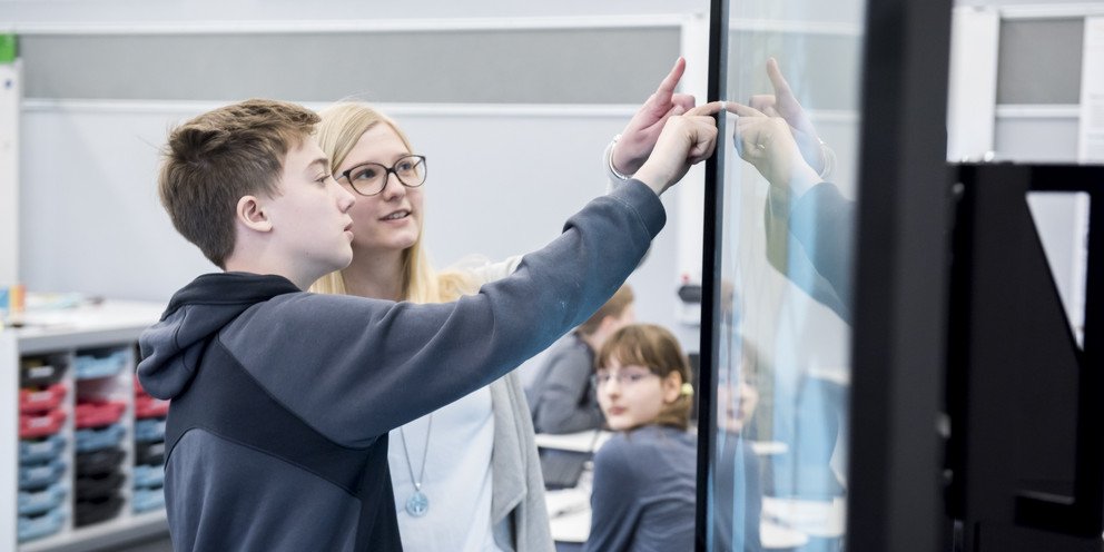 Eine Sch&uuml;ler und eine junge Studentin arbeiten gemeinsam interessiert an einem digitalen Smartboard.