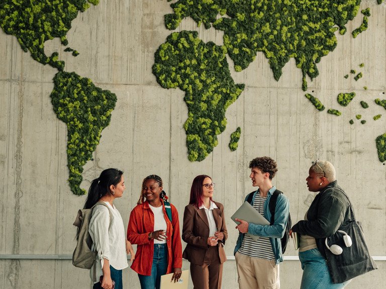 Zu sehen ist eine Gruppe junger Erwachsener welche in einer industriellen Halle vor einer gr&uuml;n bewachsenen Karte der Welt stehen. Die Personen unterhalten sich angeregt mit einem freundlichen Gesichtsabdruck. Sie stammen von unterschiedlichen kulturellen Hintergr&uuml;nden ab.