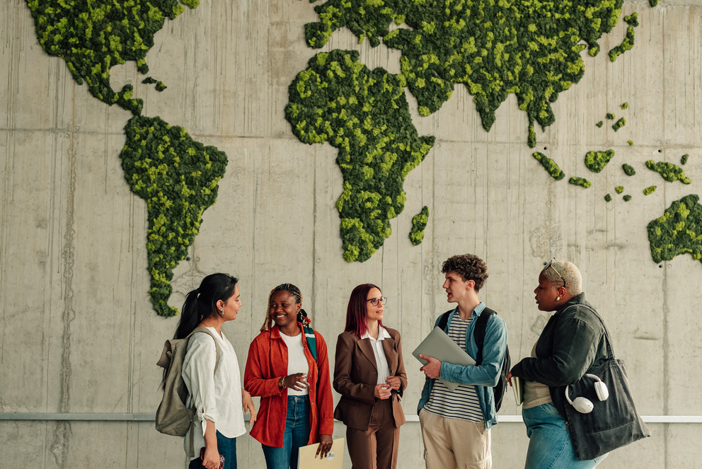 Zu sehen ist eine Gruppe junger Erwachsener welche in einer industriellen Halle vor einer gr&uuml;n bewachsenen Karte der Welt stehen. Die Personen unterhalten sich angeregt mit einem freundlichen Gesichtsabdruck. Sie stammen von unterschiedlichen kulturellen Hintergr&uuml;nden ab.