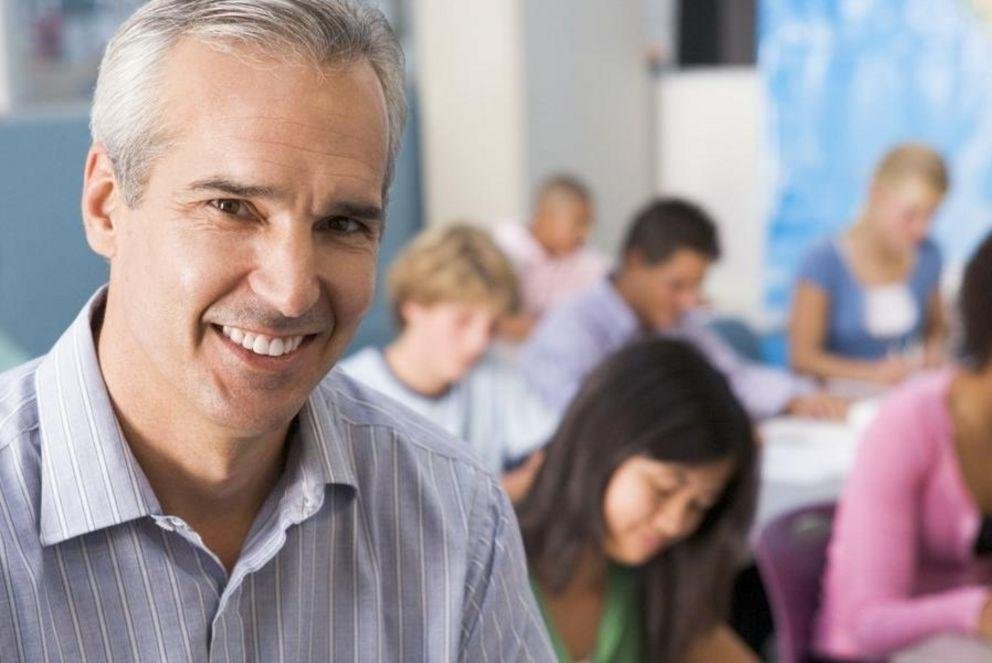 [Translate to Englisch:] Ein Lehrer steht im Klassenzimmer vor seinen Schülern und guckt in die Kamera.