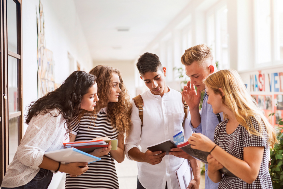 Drei Studentinnen und zwei Studenten schauen gemeinsam in ein Heft, das einer der Studenten in der Hand h&auml;lt