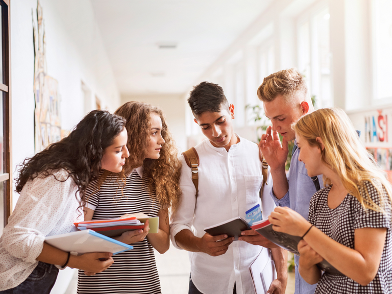 Drei Studentinnen und zwei Studenten schauen gemeinsam in ein Heft, das einer der Studenten in der Hand h&auml;lt