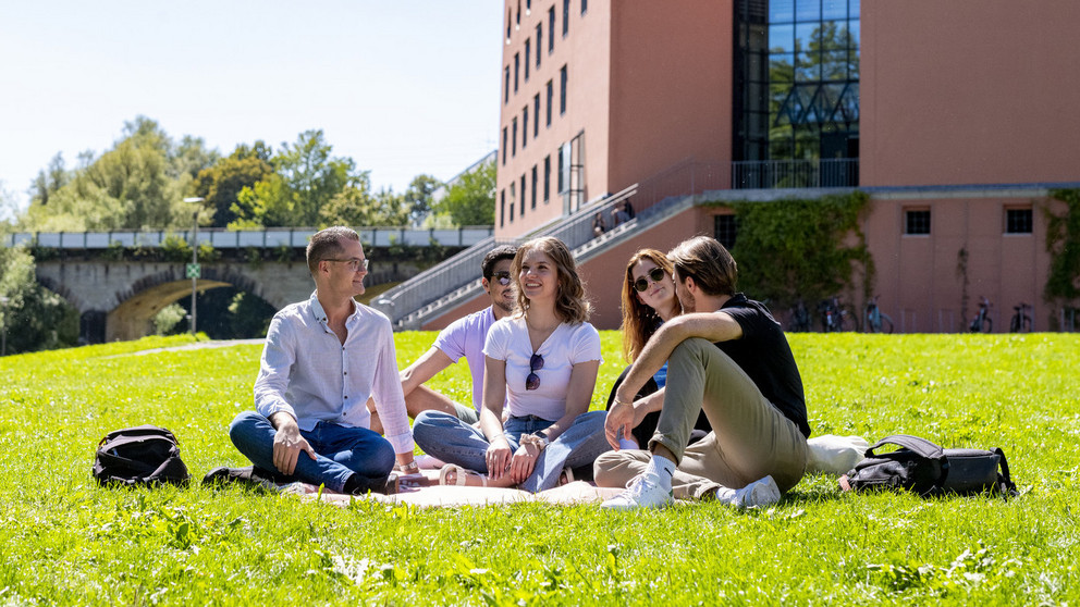 F&uuml;nf Studierende sitzen auf der Wiese vor dem Philosophicum-Geb&auml;ude der Uni Passau.