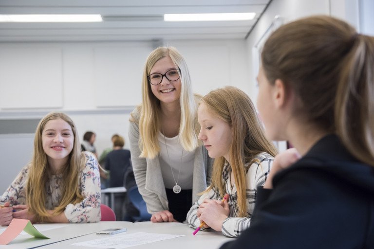 Eine Studenetin arbeitet gemeinsam mit Schlern an einem Projekt. Sie lchelt freundlich in die Kamera whrend die Schlerinnen aufmerksam weiter arbeiten.