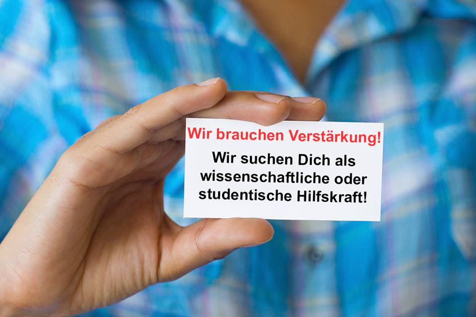 Ein K&auml;rtchen mit der Aufschrift "Wir brauchen Verst&auml;rkung! Wir suchen dich als studentische oder wissenschaftliche Hilfskraft" wird von einem Mann in die Kamera gehalten.