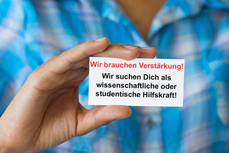 Ein Kärtchen mit der Aufschrift "Wir brauchen Verstärkung! Wir suchen dich als studentische oder wissenschaftliche Hilfskraft" wird von einem Mann in die Kamera gehalten.